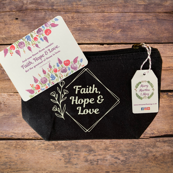 A Mary, Martha & Meg navy, organic cotton zip pouch featuring the word ‘Faith, Hope & Love,’ inside a simple design of tilted squares and flowers on the lefthand side. The design has been hand-printed. A Mary, Martha & Meg tag hangs from the zip. A small card is included with the verse, "And now these three remain: faith, hope & love, but the greatest of these is love." 1 Corinthians 13:13. It is framed beautifully by watercolour florals. Pictured on a wooden surface.