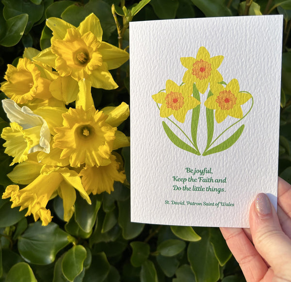 A beautifully textured eye-catching card of three golden daffodils featuring the last words of David, Patron Saint of Wales, “Be Joyful. Keep the Faith. Do the little things.” It is a lovely reminder to serve each other in the little things we do, no matter how small. This Mary, Martha & Meg card is pictured held in a hand, next to golden daffodils.