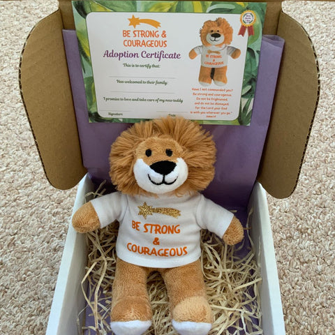 A Mary, Martha & Meg plush toy lion is seated in a box. He wears a white t-shirt with the hand-printed word, "Be strong and courageous." Above the orange text is a gold, glitter star. His adoption certificate is placed above his head.