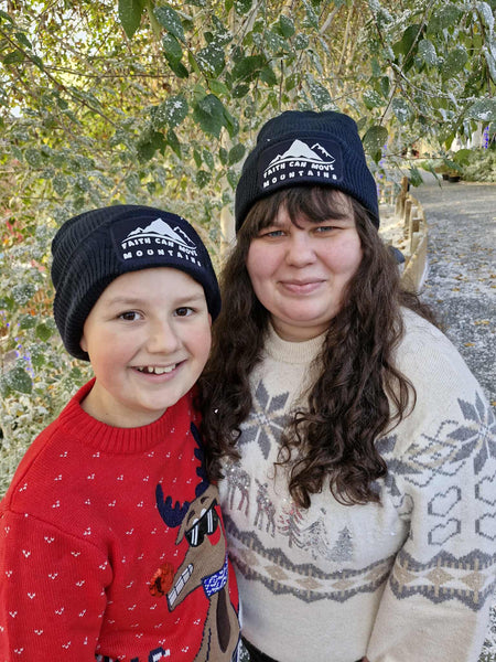 A child and a young woman in an outdoor, snowy setting. Both are wearing Christmas jumpers, one red and one cream with glitter stars. They both wear a Mary, Martha & Meg French navy beanie with white text, ‘Faith can move mountains,’ and a simple white mountain design. A warm, practical beanie suitable for children and adults alike.