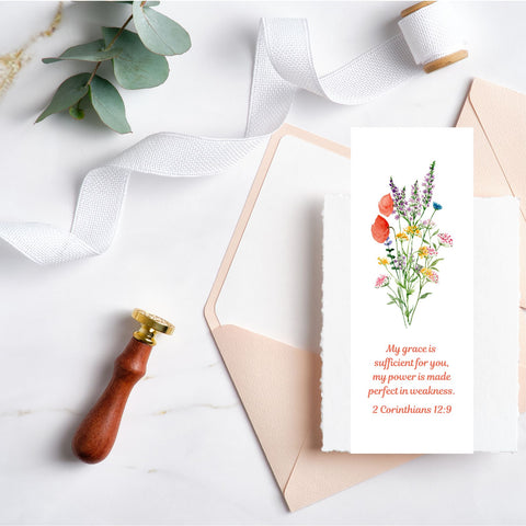 A beautiful Mary, Martha & Meg bookmark featuring a pretty wildflower floral bouquet. Underneath this is the encouraging Bible verse, “My grace is sufficient for you, my power is made perfect in weakness.” 2 Corinthians 12:9. The bookmark is pictured on a white background, to the right of carefully placed stationary items, including a wax seal stamp, green eucalyptus leaves, white cotton binding and peach envelopes. A lovely bookmark to encourage you or to give to a friend going through difficult time