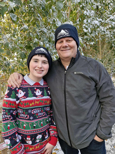A photograph of two people in an outdoor, snowy setting. Both are wearing a Mary, Martha & Meg French navy beanie featuring the text ‘Faith can move mountains.’ The child is wearing a festive Christmas jumper, whilst the man is wearing a black jacket. The beanie is a great practical gift for children and adults alike. 
