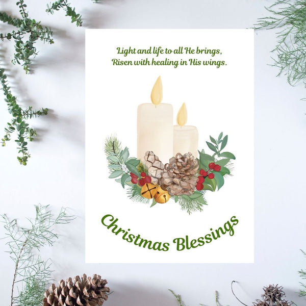 A white Mary, Martha & Meg Christmas Greeting card featuring two lit candles resting on a Christmas wreath of holly berries, pine cones, Christmas bells and foliage. The words, "Light and life to all He brings, Risen with healing in His wings" are to be found above the candles. These are taken from the carol, ‘Hark the Herald.’ The greeting, ‘Christmas Blessings’ is also included. The white card is photographed on a background of delicate green foliage and pine cones.