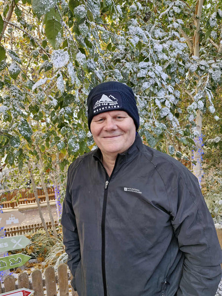 A smiling man wearing a black jacket and a French navy beanie, standing against a snowy greenery background. This Mary, Martha & Meg beanie has the encouraging text, ‘Faith can move mountains,’ in white with an accompanying white mountain design. A practical Christian gift for children and adults alike.