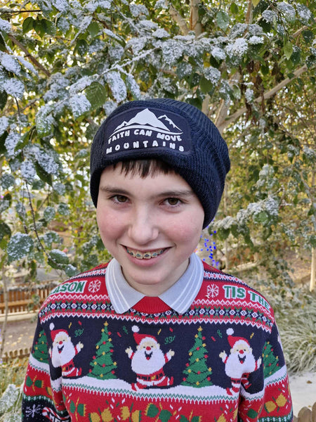A photograph of a teenager wearing a festive, Christmas-themed jumper standing in a snowy outdoor setting. He is wearing a beanie featuring the words, ‘Faith can move mountains.’ The white text and minimalist mountain design stands boldly against the classic French navy of the beanie. A lovely, practical gift for outdoor wear.