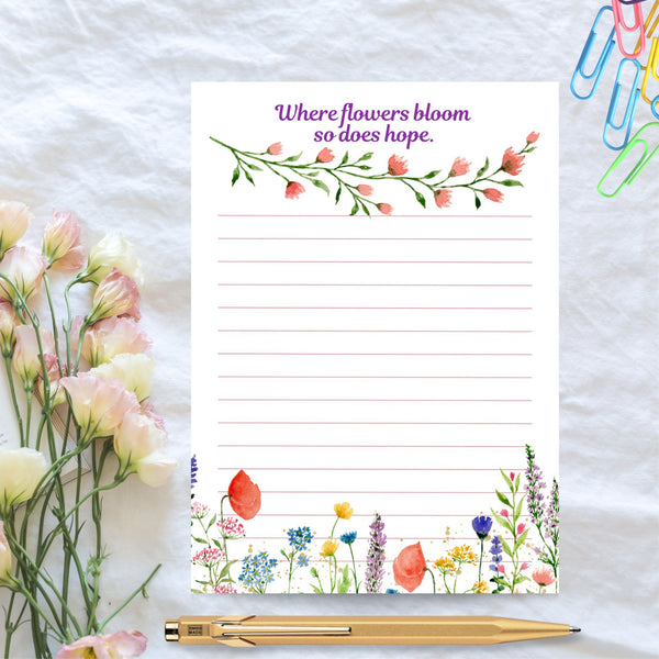 A lined notpad with 'Where flowers bloom so does hope' written at the top of the page . A beautiful curved stem of pink blooms and green leaves sits underneath the text. Pretty wildflowers can be seen at the bottom of the page. This Mary, Martha & Meg notepad is pictured on white cloth. A gold pen, co,loured paper clips and a bouquet of flowers is also pictured. A lovely gift for stationary lovers.