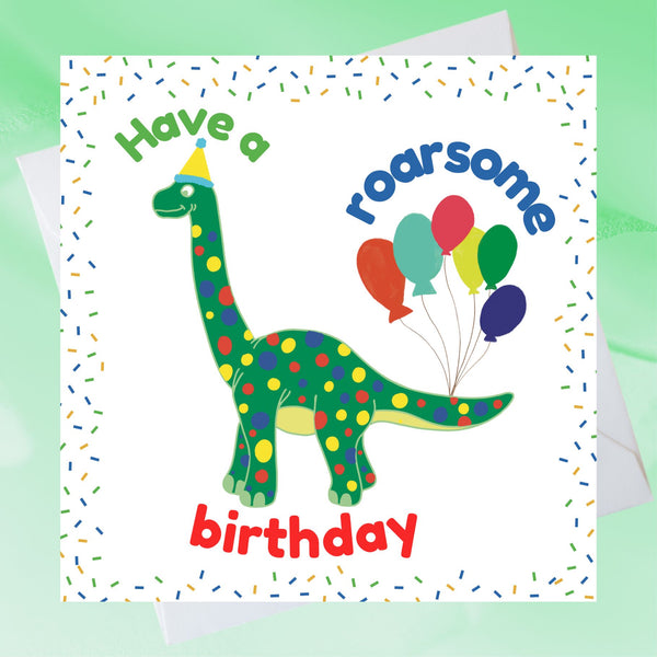 A Mary Martha & Meg child’s birthday card featuring a friendly green dotty dinosaur with party balloons tied to its tail. The text reads, ‘Have a roarsome birthday.’ The crisp white card has a colourful confetti border. A white envelope peeps out behind the card. The card is placed on a pale green background. 
