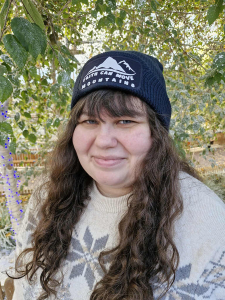 A photograph of a beanie featuring the words, ‘Faith can move mountains.’ The white text and minimalist mountain design stands boldly against the classic French navy of the beanie. This Mary, Martha & Meg beanie is worn by designer Meg, who is wearing a cream jumper with a glitter star design. She stands in front of trees dusted with snow.