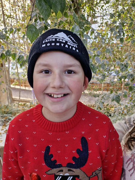 A photograph of a smiling child in a red Christmas reindeer jumper wearing a beanie featuring the words, ‘Faith can move mountains.’ The white text and minimalist mountain design stands boldly against the classic French navy of the beanie. The child is standing outdoors in front of greenery. 