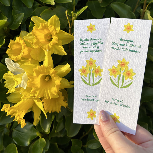 Two white bookmarks held in a hand next to bright daffodils. Both bookmarks feature the last word of St. David, one in English, one in Welsh. The Welsh reads, ‘Byddwch lawen, Cadwch y ffydd a Gwnewch y pethau bychain.’ The English reads, ‘Be Joyful, Keep the faith and Do the little things.’ Celebrate St. David with these cheerful Mary, Martha & Meg bookmarks.