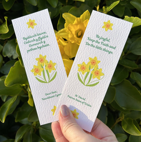 Two white bookmarks held in a hand in front of daffodils. Both bookmarks feature the last word of St. David, one in English, one in Welsh. The Welsh reads, ‘Byddwch lawen, Cadwch y ffydd a Gwnewch y pethau bychain.’ The English reads, ‘Be Joyful, Keep the faith and Do the little things.’ Celebrate St. David with these bright Mary, Martha & Meg bookmarks.