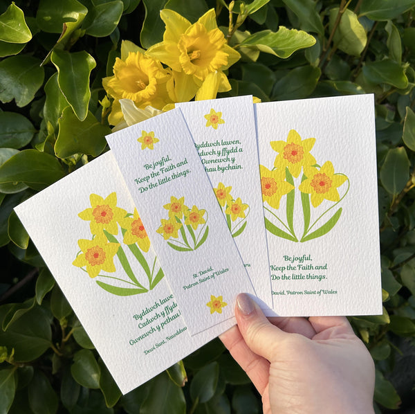 A beautiful Mary, Martha & Meg collection of daffodil designs celebrating St. David. A hand can be seen holding a card and Bookmark with English text, and another card and bookmark in Welsh. The Welsh reads, ‘Byddwch lawen, Cadwch y ffydd a Gwnewch y pethau bychain.’ The English reads, ‘Be Joyful, Keep the faith and Do the little things.’ All feature the last words of St. David, Dewi Sant. In the background are golden daffodils and leaves. 