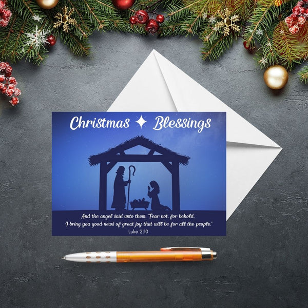 This Mary, Martha & Meg Christmas Greeting card features a Nativity scene showing the silhouettes of Mary and Joseph in a stable, worshipping baby Jesus lying in a manger. A white star is set above the stable, contrasting with the inky blue sky. The greeting at the top of the card reads, ‘Christmas Blessings.’ The text underneath the Nativity scene is from Luke 2:10, “And the angel said unto them, ‘Fear not, for behold I bring you good news of great joy that will be for all the people.”  