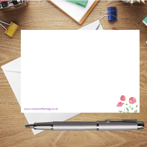 A photograph showing the reverse of a Just to Say postcard. It features a small red poppy motif in the lower right corner, as well as a small reference to the Mary, Martha & Meg website. The edge of a white envelope can be seen behind the postcard. The postcard is placed on a wooden writing surface with a silver pen beneath it and stationary items above it.