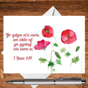 A Mary, Martha & Meg card featuring the Welsh Bible verse ‘'Yr ydym ni'n caru, am iddo ef yn gyntaf ein caru ni.' 1 Ioan 4:19. A design of three delicately hand-painted watercolour red poppies accompanies the digital text. The edges of a crisp white envelope can be seen behind the card, which rests on a wooden writing surface. A silver pen has been placed beneath the card. A perfect Welsh greetings card to remind someone how much they are cared for and loved by our Saviour.