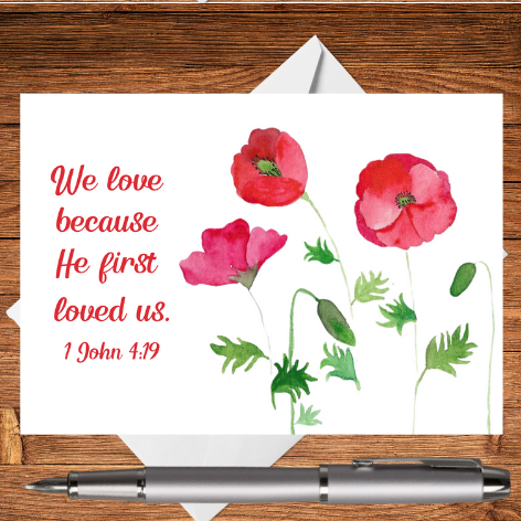 A Mary, Martha & Meg card featuring the Bible verse ‘We love because He first loved us,’ 1 John 4:19. A design of three delicately hand-painted watercolour red poppies accompanies the digital text. The edges of a crisp white envelope can be seen behind the card, which rests on a wooden writing surface. A silver pen has been placed beneath the card. A perfect card to remind someone how much they are cared for and loved by our Saviour.