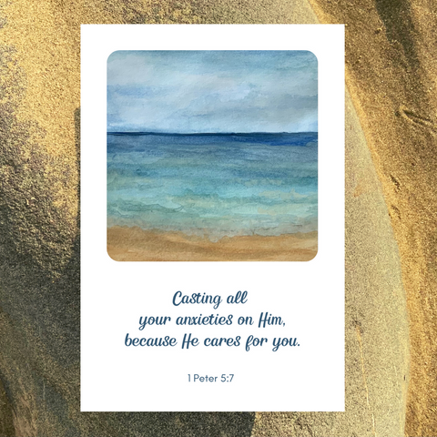 A Mary, Martha & Meg greetings card of a calming seashore. At the centre of the card is a painting of blue waves and a sandy shore, taken from an original watercolour by Meg. The Biblical text reads, “Cast all your anxieties on Him, because He cares for you.” 1 Peter 5:7 The white greetings card has been photographed against a background of golden sand.
