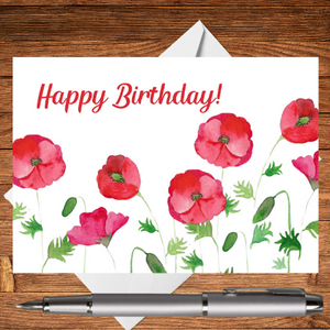 A photograph of a Mary, Martha & Meg birthday card. The card features a delicate hand-painted floral design of red poppies, complemented with Happy Birthday in digital text. The beautiful birthday card is photographed on a wooden writing surface with a white envelope peeping out behind the card. There a silver pen beneath the postcard.