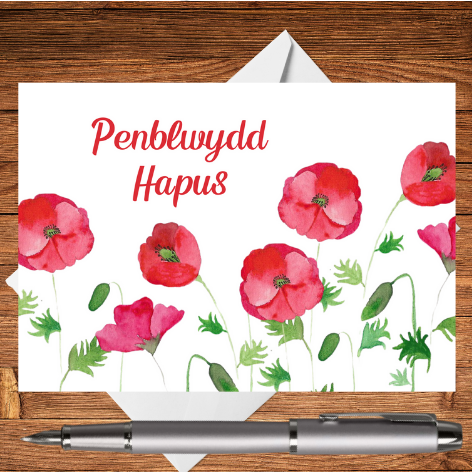 A Welsh Birthday Card featuring a classic Mary, Martha & Meg hand-painted watercolour design of delicate red poppies. These are complemented by the Welsh greeting Penblwydd Hapus (Happy Birthday). This beautiful Penblwydd Hapus card has a white envelope peeping out behind it. The Welsh birthday card is pictured on a wooden writing surface with a silver pen beneath the card.