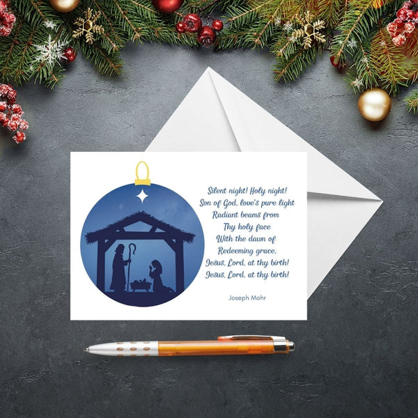 This Mary, Martha & Meg Cristmas Greetings card features a Nativity scene showing the silhouettes of Mary and Joseph in a stable, worshipping baby Jesus lying in a manger. The scene is captured inside a Christmas bauble of an inky blue sky. The text on the card features lyrics from the beautiful Christmas carol, Silent Night, written by Joseph Mohr. The card is photographed on a slate gray background with Christmas fir tree branches and festive decorations.