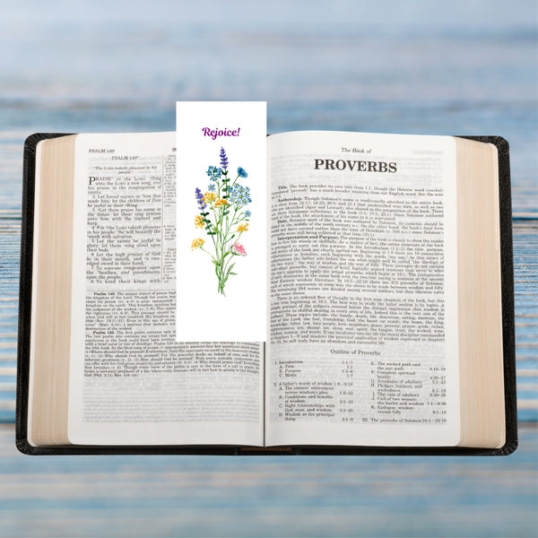 A Mary Martha & Meg white bookmark featuring the word ‘Rejoice’ above a floral bouquet of pretty wildflowers. A perfect size to pop into any book, notebook, Bible or handbag. The bookmark is photographed on an open Bible which rests on palce blue wood.