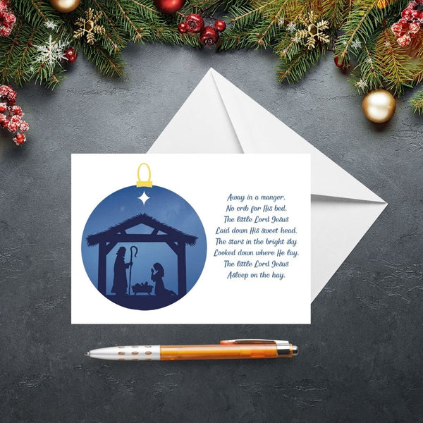 This Mary, Martha & Meg Christmas Greetings card features a Nativity scene showing the silhouettes of Mary and Joseph in a stable, worshipping baby Jesus lying in a manger. The scene is captured inside a Christmas bauble of an inky blue sky. The text on the card features lyrics from the beautiful Christmas carol, ‘Away in a Manger.’ The card is photographed on a slate gray background with Christmas fir tree branches and festive decorations.