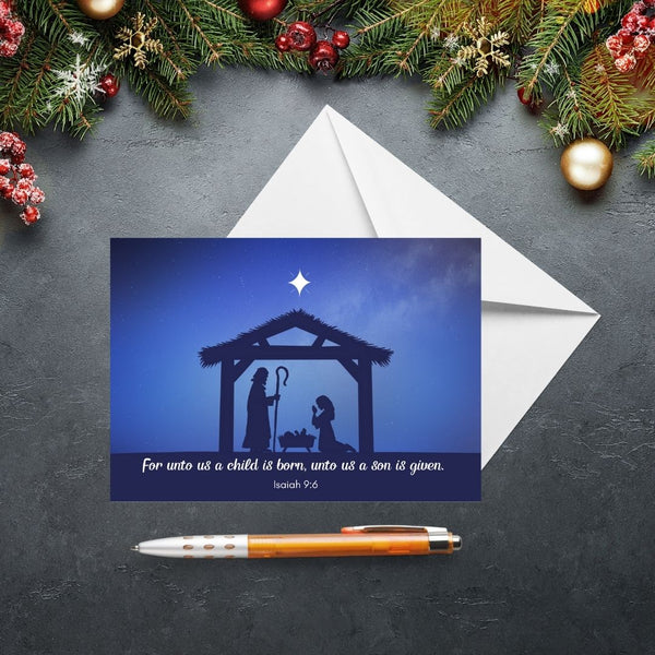 This Mary, Martha & Meg Christmas Greetings Card features a Nativity scene showing the silhouettes of Mary and Joseph in a stable, worshipping baby Jesus lying in a manger. A white star is set above the stable, contrasting with the inky blue sky. The text on the card is from Isaiah 9:6, “For unto us a child is born, unto us a Son is given.” The card is photographed on a slate gray background with Christmas fir tree branches and festive decorations.