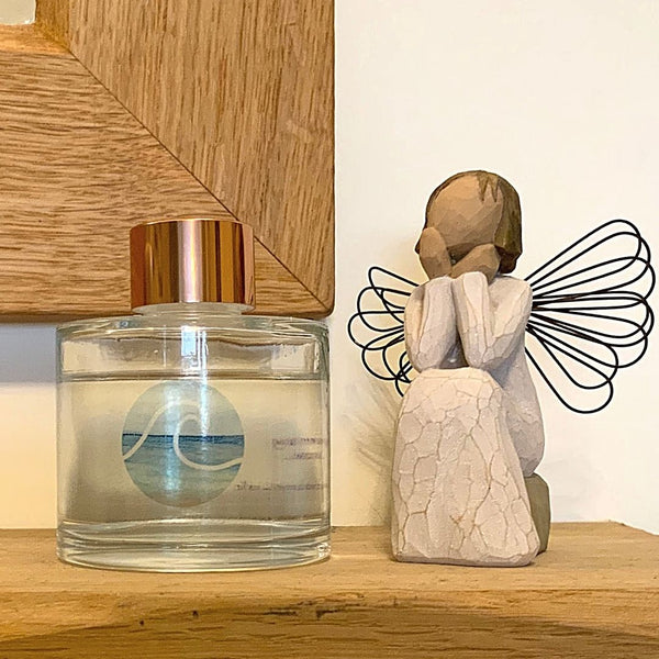 A Mary, Martha & Meg luxury reed diffuser bottle with a gold-coloured top is seen placed on a wooden mantlepiece below a wooden-framed mirror. The bottle has a simple design of a blue wave. To the right of this beautifully scented reed diffuser is a wooden sitting angel.