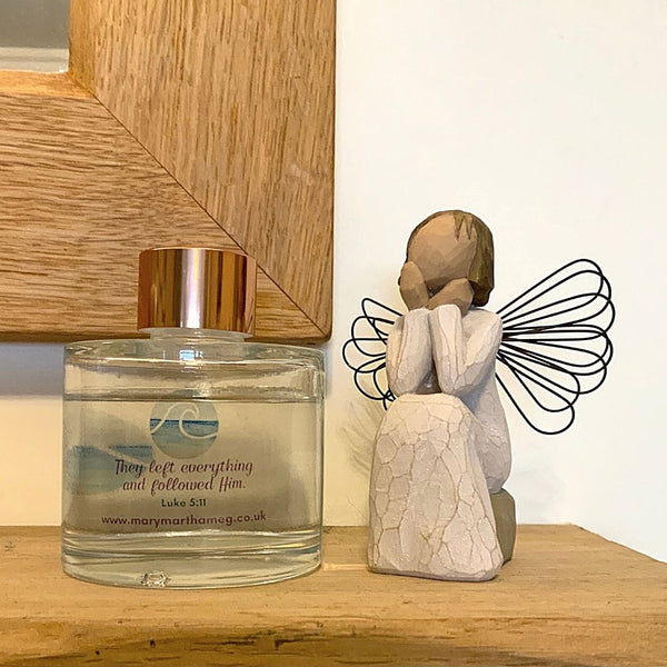 A Mary, Martha & Meg luxury reed diffuser bottle with a gold-coloured top is seen placed on a wooden mantlepiece below a wooden-framed mirror. The Bible verse which inspired this seashore fragrance, ‘They left everything and followed Him,’ Luke 5:11, is included on the bottle, as well as a reference to Mary, Martha & Meg website. To the right of the diffuser is a wooden sitting angel.