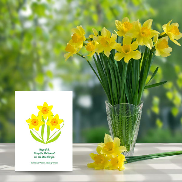 A bright, eye-catching card of three golden daffodils features the last words of David, Patron Saint of Wales, “Be Joyful. Keep the Faith. Do the little things.” It is a lovely reminder to serve each other in the little things we do, no matter how small. This Mary, Martha & Meg card is pictured next to a vase of golden daffodils, with a small bunch of daffodils lying on the table next to the vase. 