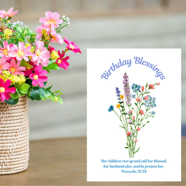 This Mary, Martha & Meg card features a simple, delicate floral bouquet, above which are written the words, ‘Birthday Blessings.’ The Bible verse written underneath the flowers is the well-known verse from Proverbs 31:28, “Her children rise up and call her blessed; her husband also, and he praises her.” The white A6 card is pictured on a pale brown shelf next to a vase of pretty pink flowers.