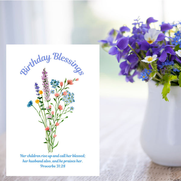 This Mary, Martha & Meg card features a simple, delicate floral bouquet, above which are written the words, ‘Birthday Blessings.’ The Bible verse written underneath the flowers is the well-known verse from Proverbs 31:28, “Her children rise up and call her blessed; her husband also, and he praises her.” The white A6 card is pictured on a cream tablecloth next to a white jug of blue, white and purple flowers.