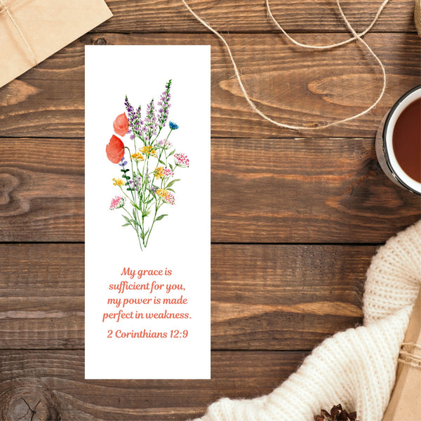 A beautiful Mary, Martha & Meg bookmark featuring a pretty wildflower floral bouquet. Underneath the floral design is the encouraging Bible verse, “My grace is sufficient for you, my power is made perfect in weakness.” 2 Corinthians 12:9. The bookmark is pictured on a brown wooden table. In the top left corner the edge of a brown parcel can be seen. In the top right corner there is some loose string. A cup of coffee and a cream jumper can be seen on the right side of the photo.