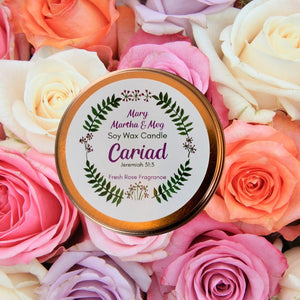 This photograph shows the lid of the Mary, Martha & Meg soy wax candle named ‘Cariad’ (Welsh for ‘Love’). This beautiful natural wax candle has a fresh rose fragrance. There is a small reference to Jeremiah 31:3, which refers to the inspiration for the candle. The text is centred inside the Mary, Martha & Meg logo wreath of green leaves and tiny purple flowers. The rose gold rim of the candle lid can be seen behind the white circular label. The lid is pictured resting on beautiful rose blooms.