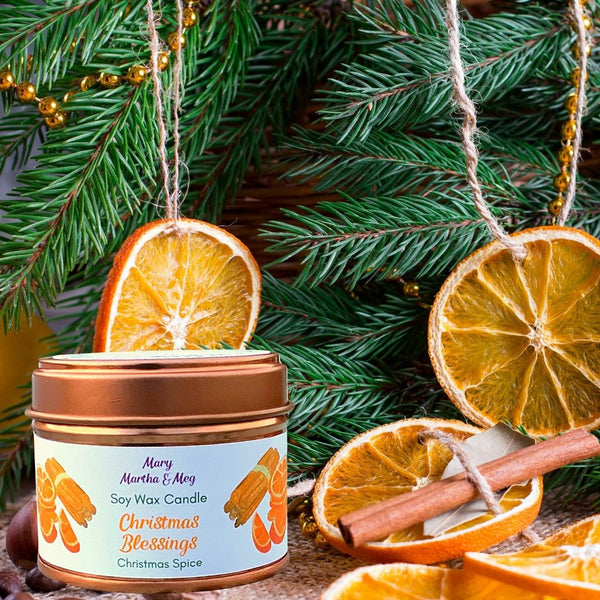 A Mary, Martha & Meg Christmas Blessing soy wax candle in a 'rose gold' tin. The candle fragrance is Christmas Spice, a delightful seasonal blend of spices and fruit associated with Christmas. The background of the photo shows Christmas tree pine leave branches decorated with hanging slices of dried orange slices tied string and cinnamon sticks.