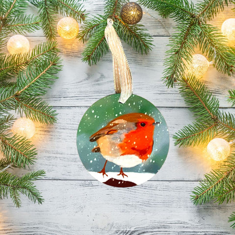 A photograph of a Mary, Martha & Meg pretty hanging decoration featuring a traditional winter robin perched on a snowy branch. The decoration is hung by a silver ribbon, ready to be placed on your Christmas tree, popped in with a Christmas card, or even added to a Christmas bouquet of flowers. The decoration is pictured on a pale wooden background and is surrounded by warm Christmas lights on pine branches.
