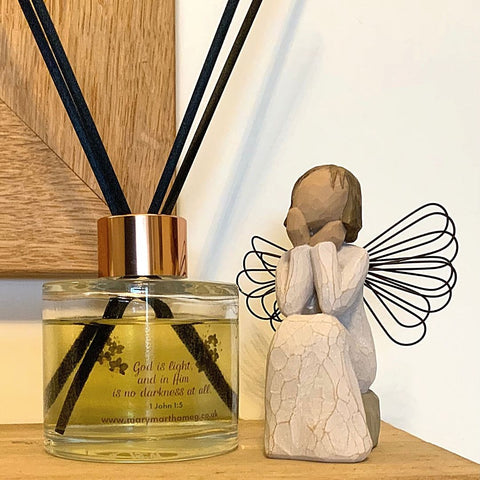 A Mary, Martha & Meg luxury reed diffuser bottle with a rose gold coloured top is seen placed on a wooden mantlepiece below a wooden-framed mirror. The Bible verse which inspired this Black Orchid ‘Hope’ fragrance can be seen, “God is Light and in Him is no darkness at all.” 1 John 1:5. Below this is a reference to Mary, Martha & Meg website. To the right of the diffuser is a wooden sitting angel. 