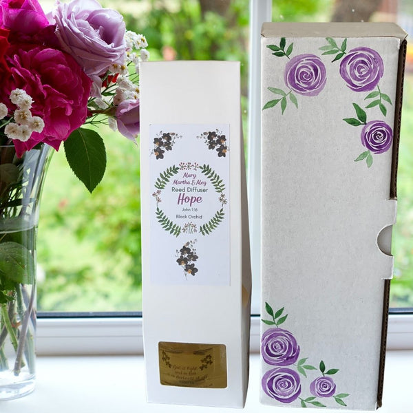 A Mary, Martha & Meg reed diffuser box is pictured on a windowsill with a garden view. The box shows the Mary, Martha & Meg logo, inside of which can be seen the name of the diffuser, ‘Hope’ as well as the name of the fragrance, ‘Black Orchid.’ A glimpse of the diffuser bottle can be seen through the rectangular cut-out in the box.  To the right of the box is a hand-painted box of purple roses. A vase of roses can be seen to the left of the diffuser. 