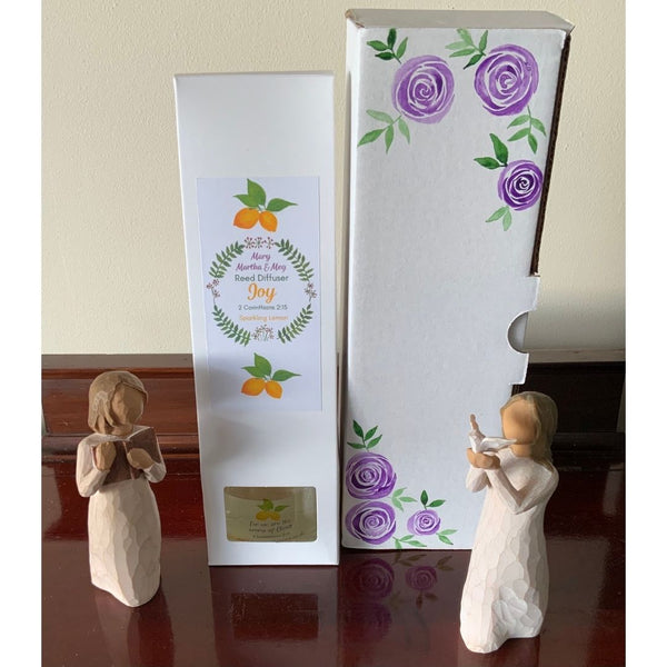 A Mary, Martha and Meg ‘Joy’ Reed Diffuser in its white packaging. The front of the white box has the Mary, Martha & Meg logo. Written text includes the name of the diffuser ‘Joy,’ the Bible verse and reference which inspired this fragrance, and the name of the fragrance, ‘Sparkling Lemon.’ There is an illustration of lemons above and below the logo design. Next to the packaging is a hand-painted box of purple roses. The reed diffuser packaging is placed between two wooden figurines.