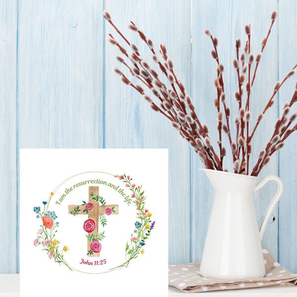 A Mary, Martha & Meg Easter card pictured on a white shelf next to a white jug of Spring buds, with a pale blue wooden background. At the centre of this white, square card is a cross framed by a delicate wreath of pretty flowers. A beautiful card to give to a friend to share the joy of Easter.