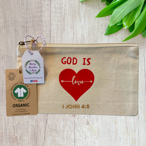 A Mary, Martha & Meg organic, natural, canvas zip pouch featuring the treasured Bible verse, ‘God is Love, 1 John 4:8.’ The word ‘love’ is featured inside a red heart and is designed as an arrow. The Bible reference, 1 John 4:8, is in gold glitter. The design has been hand-printed. A Mary, Martha & Meg tag hangs from the zip, as well as a label showing the organic credentials of the zip pouch. The zip pouch is pictured on a wooden table with green leaves in the top right corner. 