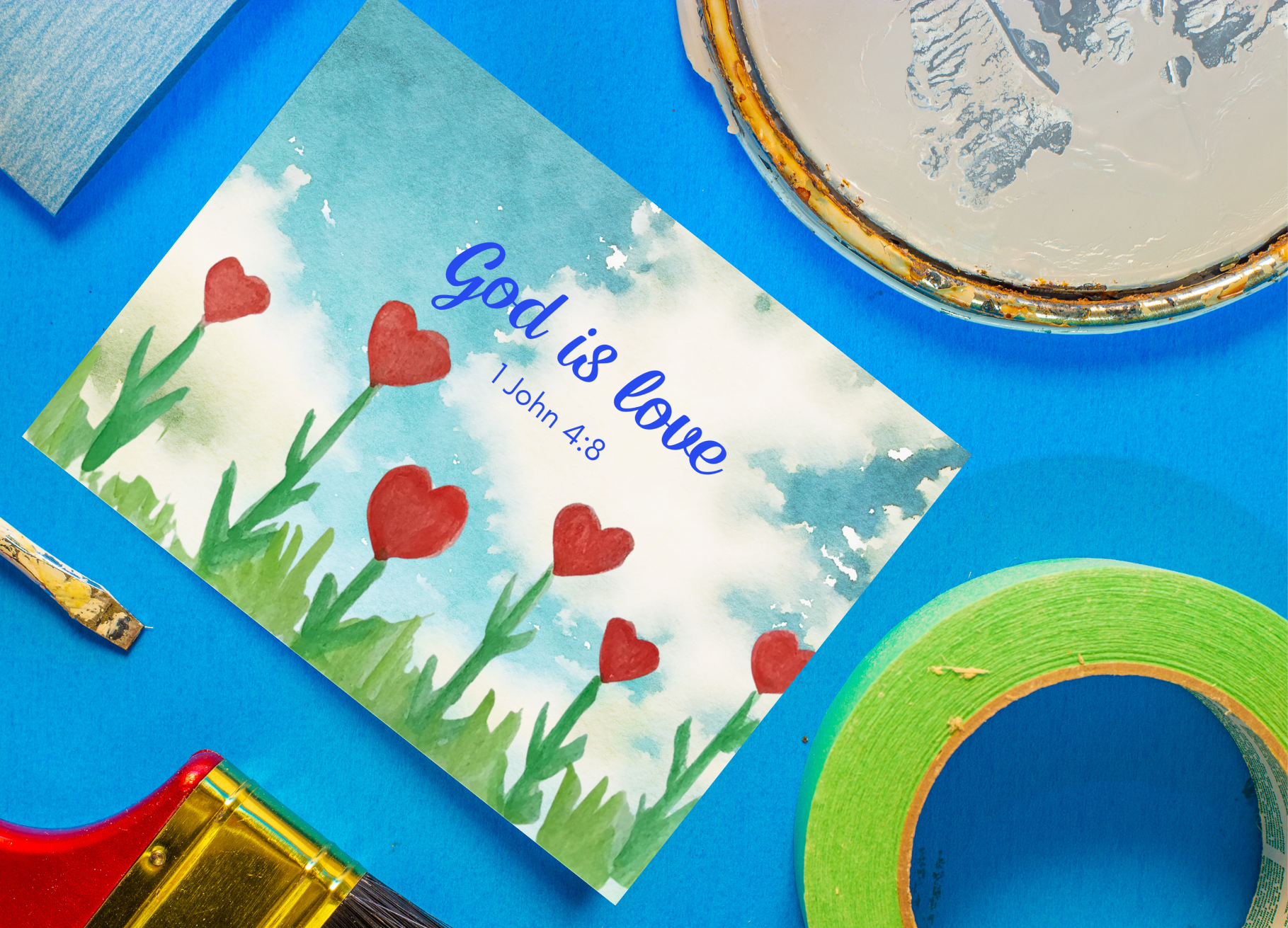 A photograph of a postcard with the words ‘God is Love, 1 John 4:8.’ The design is of red heart flowers growing between blades of green grass. Purple and yellow butterflies flutter in the blue sky. This watercolour has been hand-painted by a young child. The background of the photographs is blue. There are paint items surrounding the photo (card, a paint tub lid, green tape, a red handled brush and a smaller paintbrush).