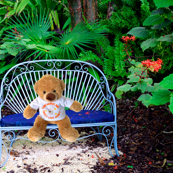 A photo of a Mary, Martha & Meg plush Honey Bear sitting on a miniature blue bench in a woodland setting of green leaves, orange flowers, tree bark. This beautifully soft bear wears a white t-shirt with the hand-printed orange text, "Kind words are sweet like honey." Honey Bear is a wonderful reminder of God's encouragement to be kind and show love to others.  