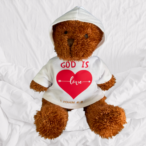 This Mary, Martha & Meg beautiful bear is a perfect reminder of God’s love for us. James is a soft plush bear with brown fur. He wears a soft white hoodie which has been carefully hand-printed with the words ‘God is Love,’ 1 John 4:8. A white arrow runs through the word ‘love,’ which is in the centre of a red heart. The Bible reference, 1 John 4:8, is made of gold glitter. James’ beautiful facial features have been embroidered. This adorable soft plush bear is photographed on a white cotton sheet. 
