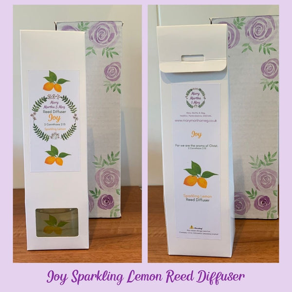 Two photographs showing the front and reverse of the white packaging boxes for Mary, Martha & Meg 'Joy' reed diffusers. Behind these 'Sparkling Lemon' fragrance diffusers are white packaging boxes hand-painted with purple roses and green foliage.