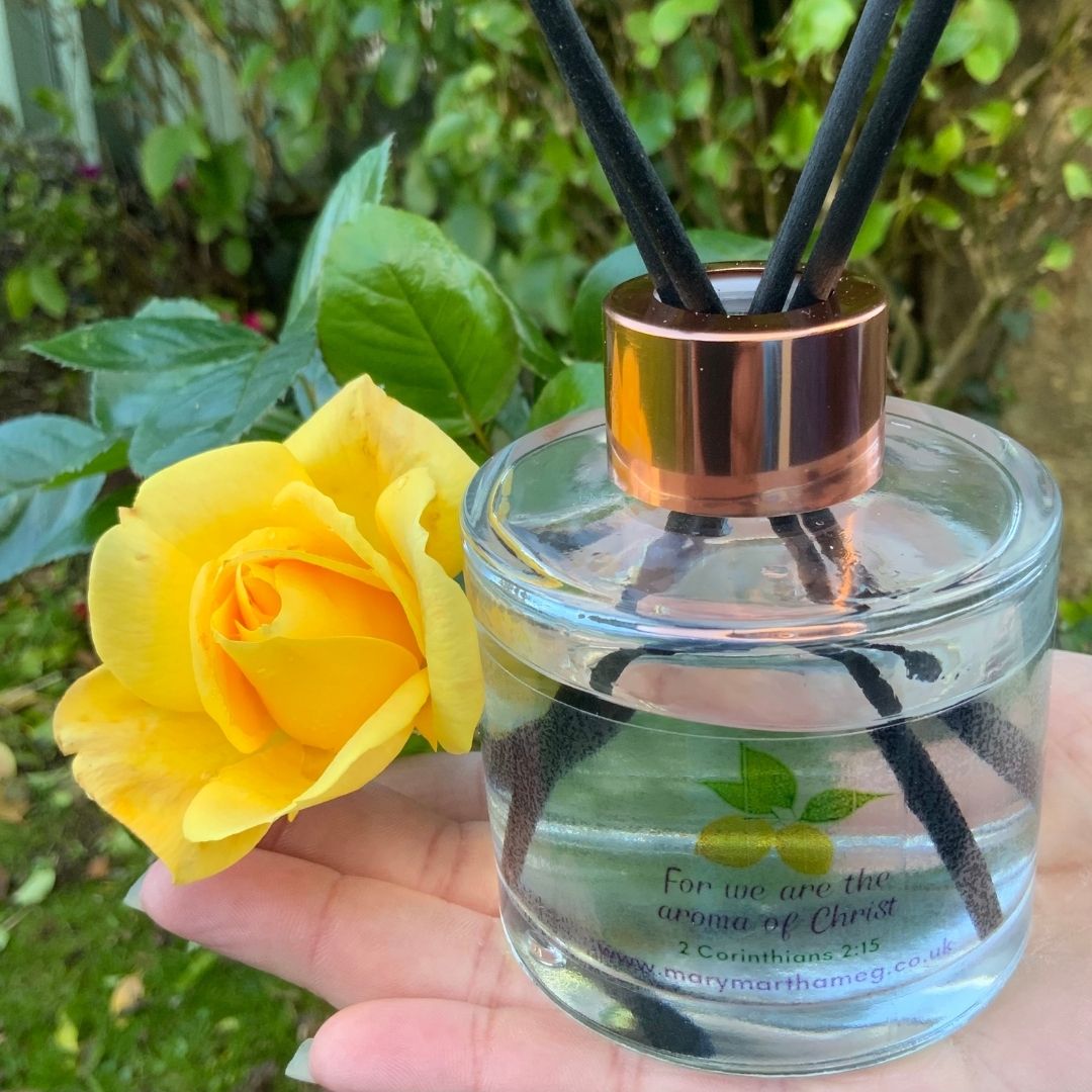 This beautiful Mary, Martha & Meg luxury reed diffuser bottle can be seen held in Meg’s hand. The fresh lemon fragrance is illustrated with two lemons on the bottle, as well as the words “For we are the aroma of Christ,” 2 Corinthians 2:15.’ In Meg’s hand can also be seen a yellow rose, with garden foliage in the background.
