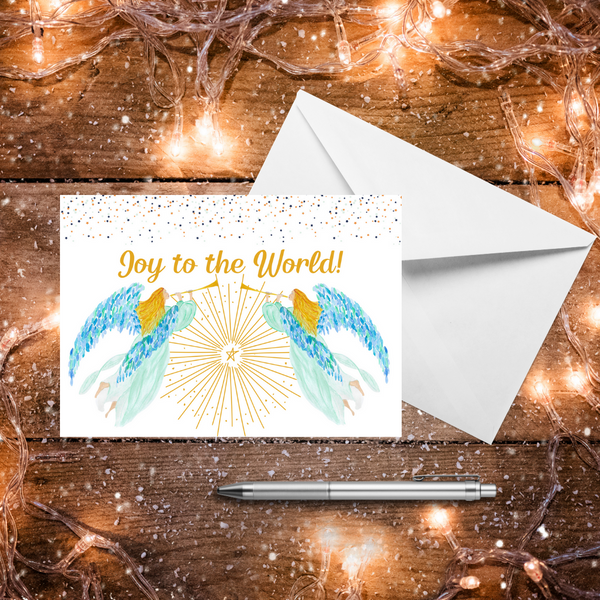 A Mary, Martha & Meg Christmas Greeting card featuring two heralding angels and a vibrant shining star between them. The golden-haired angels wear delicate mint gowns and have beautiful blue and green feathered wings. Each carries a golden trumpet. The text reads, ‘Joy to the World!’ Above this are tiny stars. A white envelope sits behind the card. The card and envelope are photographed on a wooden table with warm Christmas lights and a silver pen.