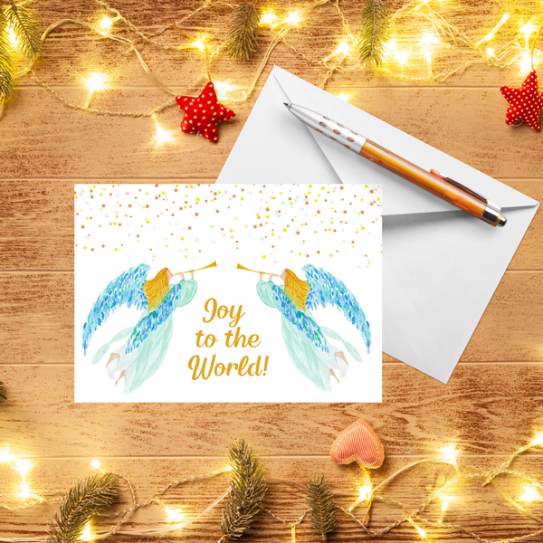 A Mary, Martha & Meg Christmas Greeting card featuring two heralding angels. The golden-haired angels wear delicate mint gowns and have beautiful blue and green feathered wings. Each carries a golden trumpet. Above them shine tiny stars. The text reads, ‘Joy to the World.’ A white envelope and pen are behind the card. These are photographed on a wooden table with warm Christmas lights and red handmade decorations. 