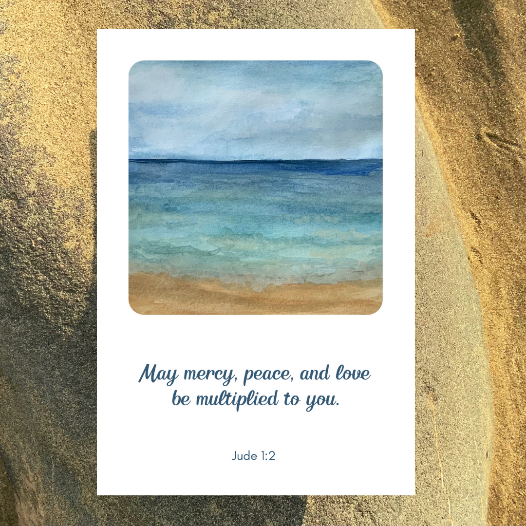 A Mary, Martha & Meg greetings card with a prayer of three blessings. The text of scripture words reads, “May mercy, peace, and love be multiplied to you.” Jude 1:2. The square painting in the centre of the white card is a print of an original watercolour painting by Meg of a seashore showing blue waves and a sandy seashore. The card is photographed on a sandy beach near Meg’s home.