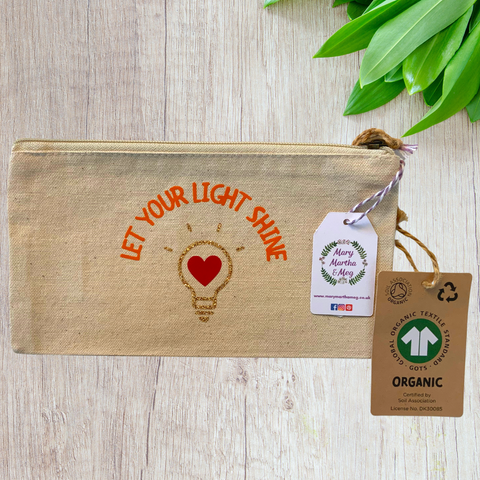 A Mary, Martha & Meg organic, natural, canvas zip pouch featuring the encouraging words ‘Let your light shine.’ Below this is a light bulb made from gold glitter vinyl. Inside the lightbulb is a red heart. The design has been hand-printed. A Mary, Martha & Meg tag hangs from the zip, as well as a label showing the organic credentials of the zip pouch. The zip pouch is pictured on a wooden table with green leaves in the top right corner. A great gift which can be posted through a letterbox.